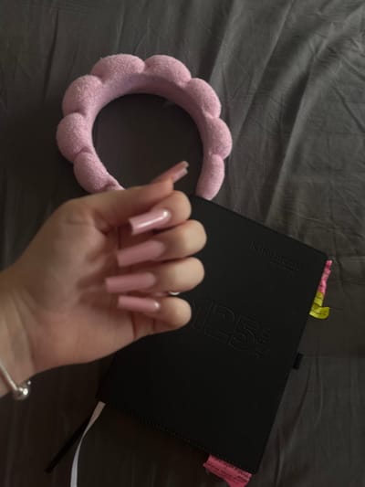 An overhead shot shows a person's hand with long, pink, square-shaped acrylic nails resting on a black notebook. The notebook has the text "Klio-Eterna 125 Jahre" embossed on its cover. A pink, fluffy headband with a scalloped edge lies just above the notebook. The scene is set against a dark gray, rumpled fabric surface. The image does not contain any information about the location in Hausach, Germany.