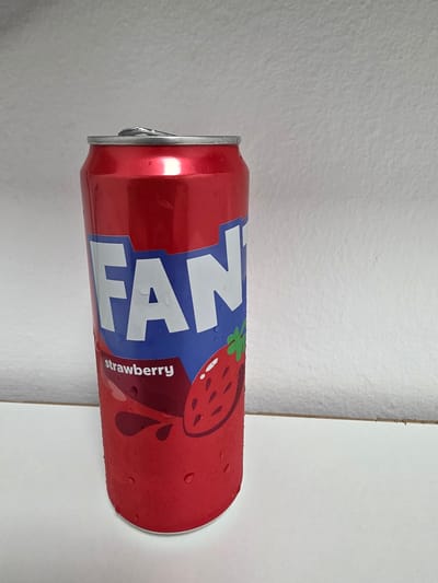 A red Fanta can, frosted with condensation, rests on a white surface. The can features the Fanta logo in blue and white, with the word "strawberry" printed in white text beneath a stylized strawberry graphic. The background is a textured, light-colored wall. The location is not specified in the image, therefore it cannot be identified as Dibba Province, Oman.