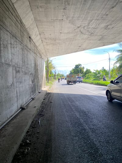 The image shows a view from under a concrete overpass on a road in Payarangkuluh, Indonesia. The overpass casts a shadow on the left side of the frame, revealing a rough, gray concrete wall with visible wiring running along it. The road surface is asphalt, with tire tracks and some debris visible along the edge.
Several vehicles are present on the road. In the distance, a light-colored truck is driving away from the viewer. Ahead of it, a white SUV is also moving forward. Closer to the foreground, on the right side of the road, the right side of a silver car is visible. A person on a motorcycle is seen further down the road, heading away from the camera.
The environment is tropical, with lush green vegetation and palm trees lining the right side of the road. A street lamp is also visible in the distance. The sky is blue with some scattered clouds. The overall scene depicts a typical day on a road in a tropical urban or semi-urban area.