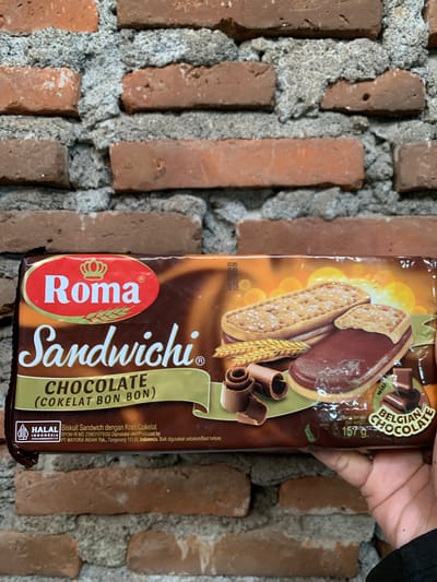 A package of Roma Sandwhichi Chocolate cookies is held in a person's hand against a brick wall. The cookies are chocolate flavored sandwich cookies with a cream filling. The package has "ROMA SANDWICHI CHOCOLATE (COKELAT BON BON)" printed on it. The cookies are depicted with a chocolate filling and a biscuit exterior. There are also chocolate curls and pieces of chocolate shown on the packaging. A "HALAL" certification is visible on the lower left side of the package, along with Indonesian text indicating it is produced by PT MAYORA INDAH Tbk. in Tangerang, Indonesia. The brick wall is composed of reddish-brown bricks with grey mortar. The background is out of focus, but appears to be an outdoor setting. There is no indication of the location in Karangkemiri, Indonesia.