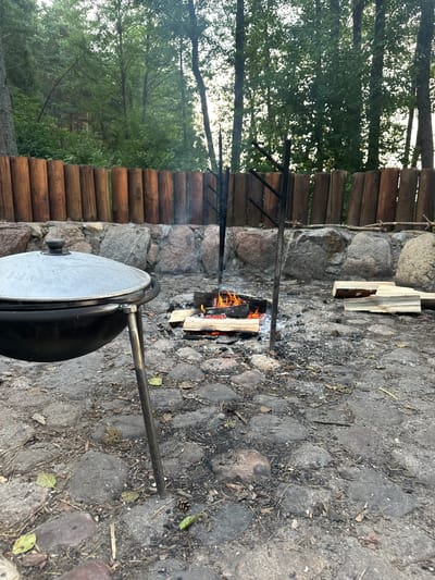 The image depicts an outdoor cooking scene in a wooded area. In the foreground, a large, dark, lidded pot is placed on a metal tripod. Adjacent to it, a small campfire burns, with logs arranged for fuel. The ground is paved with irregular stones and shows signs of ash and scattered debris like pinecones and leaves. Behind the fire pit, a low wall constructed from large, rough-hewn stones forms a boundary. Beyond this wall, a rustic fence made of vertically placed wooden logs extends across the frame. In the background, a dense forest with tall trees and green foliage is visible. The lighting suggests it is either early morning or late afternoon, with diffused light filtering through the trees. There are no people present in the image.