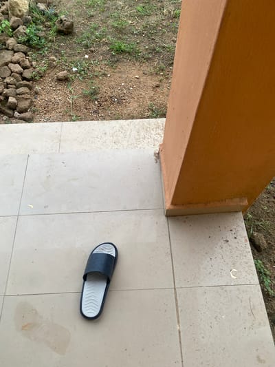 This image depicts an outdoor scene in Dumnga, Nigeria. The foreground is dominated by a tiled floor, likely a patio or walkway, that shows signs of dirt and some reddish-brown stains. A single, dark blue and white sandal rests on the tiles. To the left of the tiled area is a patch of unpaved ground with sparse green grass and small stones. A large, orange-colored beam or pillar runs diagonally across the upper left portion of the frame, casting a shadow that is not visible in the image. The ground beyond the tiles appears to be uncultivated earth with more scattered stones and dry vegetation. The lighting suggests it is daytime, though the weather conditions are not explicitly evident. The scene appears to be a residential or communal outdoor space. No people are visible, and there is no visible text.