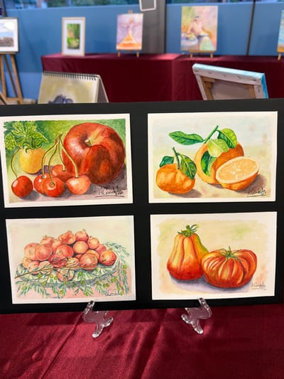 This image shows a display of watercolor paintings of fruits and vegetables, likely at an art exhibition or market. In the foreground, four small watercolor paintings are arranged on a black mat, with clear acrylic stands holding them upright. The paintings depict: a red apple with a yellow tomato and cherries; oranges, one sliced in half; a pile of pomegranates with greenery; and two gourds or pumpkins with a tomato. The backdrop reveals a display table covered in a dark red velvet cloth, with other paintings and easels visible in the blurred background, suggesting an indoor event. The style of the paintings appears to be realistic, with attention to light and shadow. Without additional context, the specific location within Elx/Elche cannot be determined.