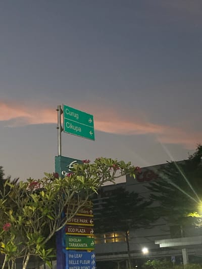 A signpost stands in the foreground, bearing directional signs for "Curug" and "Cikupa." Below these, a series of smaller signs indicate "OFFICE PARK," "ECO PLAZA," "SEKOLAH TARAKANITA," "the LEAF," and "BELLE FLEUR." A flowering tree with pink blossoms is positioned to the left of the signpost. In the background, a building with a red logo is visible, and the sky exhibits a twilight hue with streaks of pink clouds. The setting appears to be in the city of Mekar Bakti, Indonesia, during dusk.