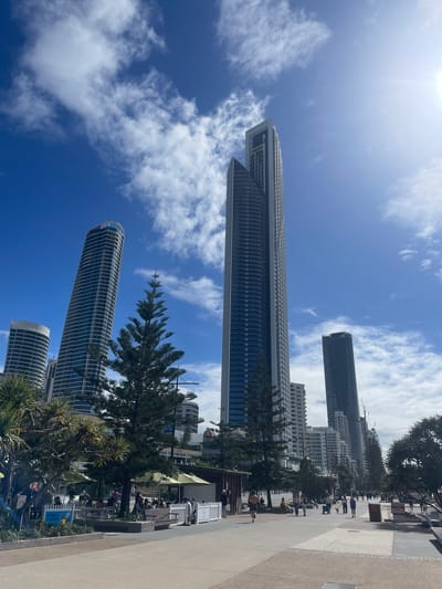 The image shows a sunny day at Surfers Paradise beach in Gold Coast, Australia. The scene features a wide, paved walkway bordering the beach, with a line of palm trees and pine trees providing shade. Several tall, modern high-rise buildings dominate the skyline, including the iconic Peppers Soul Surfers Paradise building.

On the walkway, people are strolling, walking dogs, and sitting at outdoor cafes under green umbrellas. A sign that reads "BECOME A LIFESAVER" is visible in the foreground. The sky is a vibrant blue with scattered white clouds, and the sun is shining brightly. The overall atmosphere is one of a bustling beachside urban environment.