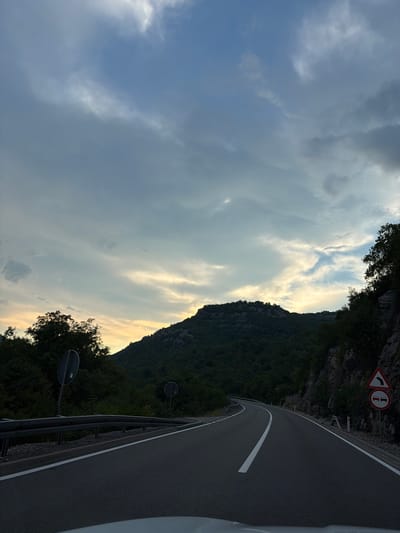 The image depicts a winding road carved into a mountainside, with a dense forest blanketing the slopes. The road, asphalted and marked with a white line, curves to the right, bordered by a protective metal barrier. Traffic signs are visible along the road: one indicates a sharp left turn and another is a circular sign with two white lines, possibly related to traffic restrictions.
The sky above is dramatic, filled with a mix of dark, heavy clouds and lighter, illuminated patches, suggesting the sun is setting or has recently set, casting a warm glow. The overall mood is serene yet imposing, with the vastness of the sky and mountains contrasting with the man-made road. The scene is likely captured in the late afternoon or early evening, given the quality of light. The location appears to be mountainous and rural, fitting the context of Gospoština, Montenegro. No people are visible in the image, only the elements of the natural landscape and the infrastructure for travel.