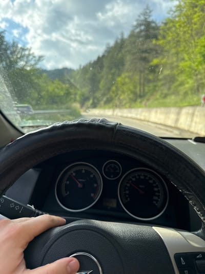 A car's interior is shown, with a view through the windshield of a road in Byala Reka, Bulgaria. The dashboard, steering wheel (with a worn cover), and instrument panel are visible. A hand is resting on the steering wheel. The exterior view features trees, sky with clouds, and a stretch of road.