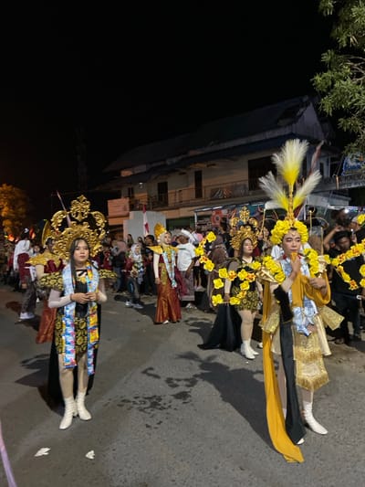 This image depicts a nighttime procession on a paved street in Kedungbanteng, Indonesia.

The scene features approximately 6-8 individuals, primarily young adults, participating in a cultural event or parade. These individuals are dressed in elaborate, ornate costumes characterized by large, golden, sculptural headpieces. Some headpieces are further embellished with white and yellow feathers, notably on the individual to the far right. The costumes incorporate various fabrics, including gold, black, maroon, and yellow, often with intricate patterns and adorned with garlands of artificial yellow flowers. Several participants, including the two prominent figures in the foreground, have multiple small, packaged items (resembling consumer product sachets) attached to sashes draped over their shoulders or integrated into their attire. Footwear for the foreground individuals consists of white ankle boots. Their expressions are generally focused or neutral as they move along the street.

In the background, a crowd of spectators and additional participants in casual attire lines both sides of the street, observing the procession.

The environment consists of a dark asphalt road, showing some minor stains and two small, crumpled white objects on the surface in the foreground. On the right side of the street, a two-story building with a dark blue corrugated roof, white walls, and a balcony is visible. An Indonesian flag (red and white) is attached to the building. A partially obscured sign on the building reads "DEEA KALIDA..." and "RT 12 RW0...". Dark foliage is visible on the far right and left edges of the frame. The sky is completely dark, indicating nighttime, with the scene illuminated by unseen artificial light sources, casting shadows from the participants onto the street.