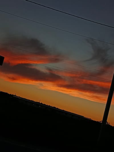 A silhouetted landscape against a sunset sky, likely located in the city of Savino, Russia, is depicted. The sky is a gradient of deep blue at the top, transitioning to fiery orange and red hues closer to the horizon, indicating a sunset. Wispy clouds are illuminated by the setting sun, creating streaks of vibrant color across the sky. Power lines stretch horizontally across the upper portion of the frame. The lower part of the image is dominated by a dark silhouette of the land, which appears to be a hillside or elevated ground with some indistinct structures or vegetation along its edge. A tall, thin pole, possibly a utility pole or lamppost, stands vertically on the right side of the image, also silhouetted against the sky. The overall impression is a serene, albeit slightly industrial or rural, evening scene.
