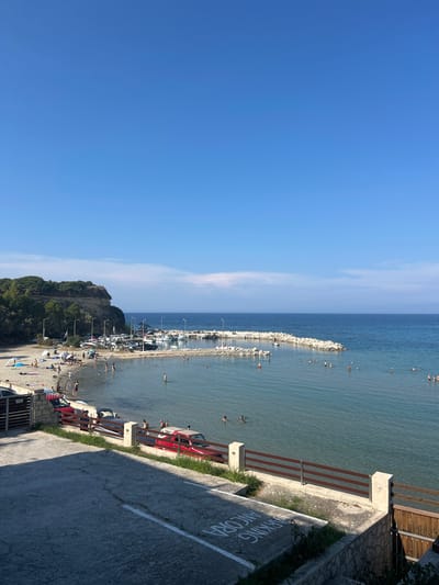This media file shows a lively beach scene on a sunny day. The image is oriented vertically, with the deep blue sky occupying the left half and the sea and shore filling the right. Along the coastline, a pier or breakwater extends into the calm, clear blue water, lined with rocks and dotted with people swimming and enjoying the sea.

The beach itself is a sandy expanse filled with numerous people. Some are sunbathing on towels, while others are walking along the shore or engaging in water activities. There are also several parked cars, some red and others white, situated near the beach, suggesting a parking area. Green trees and a rocky hill rise behind the beach, adding to the natural landscape.

The overall atmosphere is one of leisure and recreation. The bright sunlight and clear sky indicate pleasant weather, likely during midday or early afternoon. The presence of many people enjoying the water and beach suggests a popular tourist or local spot.

Visible text on the pavement in the foreground reads "PARKING ANCORA" in white stencil lettering, indicating a parking area. The location context provided places this scene in Arkadii Municipal Unit, Greece.