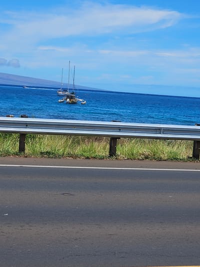 The image depicts a scenic coastal highway scene in Lahaina, Hawaii. The viewpoint is from the road, looking out towards the ocean. In the foreground, a dark asphalt road with white and yellow lane markings stretches horizontally. To the left of the road, a metal guardrail separates it from a grassy embankment.
Beyond the embankment, the vibrant blue ocean occupies the majority of the frame. Several sailboats are visible on the water, some anchored and others seemingly in motion. One prominent sailboat, with multiple masts, is closer to shore. The sky above is a clear, bright blue with scattered white clouds, suggesting a pleasant day. The overall impression is one of a serene coastal environment, typical of a Hawaiian roadside view. There is no visible text in the image. The time of day appears to be daytime, with ample sunlight illuminating the scene.