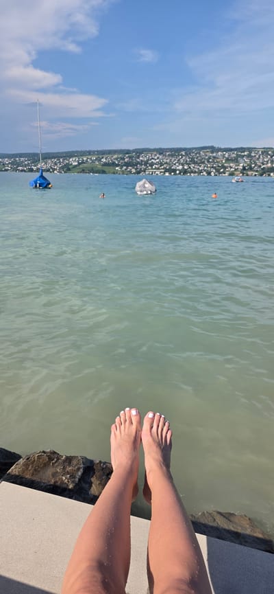 In Oberrieden, Switzerland, a first-person perspective shot shows a person's bare legs and feet dangling over a stone embankment and into a body of water. The feet are clean with white nail polish. The water is calm and has a greenish hue, with ripples on the surface. In the background, a town with white buildings is visible on the shore, nestled against green hills under a blue sky with scattered clouds. A sailboat with a blue sail and another covered boat are anchored in the water. A few people are swimming in the distance. The overall scene depicts a sunny day at a lake or large body of water.