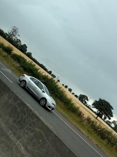 The image depicts a silver sedan in motion on a road that appears to be a rural highway or country lane, possibly in Ballycanew, Ireland. The car is traveling away from the camera and slightly to the left, with its wheels blurred due to motion. The road surface is dark asphalt with a yellow line marking the center and white lines marking the edges. To the right of the road, a field of cut or ripening grain, likely wheat or barley, stretches into the distance. This field is bordered by dense green shrubbery and scattered trees, including a mature oak tree visible further down the field. Overhead, the sky is overcast with a pale grey, cloudy hue. In the background, power lines and pylons are visible, suggesting proximity to a rural infrastructure network. The foreground shows a blurred section of what could be a roadside barrier or the edge of the vehicle from which the photo was taken, also angled. The overall scene conveys a sense of travel through a rural landscape under a cloudy sky.