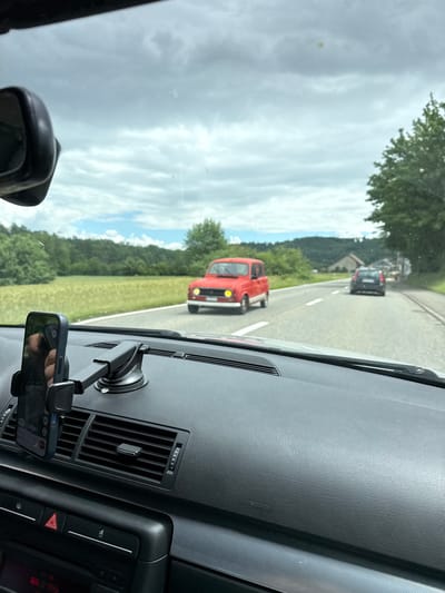 The image depicts a daytime road scene in a rural or semi-rural area, viewed from inside a car in Zurzach, Switzerland. The sky is bright but cloudy, overlooking expansive green fields, dense trees, and distant hills. On the asphalt road ahead, marked with white lines, a distinctive red vintage car (possibly a Renault 4) is directly in front, followed by a darker modern vehicle further down the road. A building is faintly visible behind the second car amidst the trees.

The foreground shows the interior of the car, featuring a dark grey, textured dashboard. An air vent is present on the right, and a phone holder, secured by a suction cup, clutches a dark smartphone. A hand is visible holding the phone. A rear-view mirror is partially seen on the left. The dashboard prominently displays an "AIRBAG" emblem on the passenger side. A control panel to the right includes a red triangular hazard warning button. The scene portrays a calm journey through scenic countryside under an overcast sky.