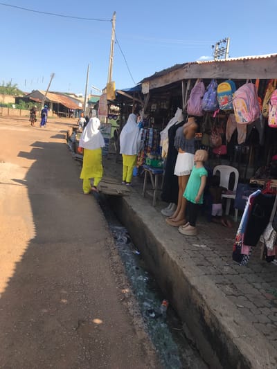 This street-level photograph, taken in Mandate Estate, Ilorin, Nigeria, captures a vibrant outdoor market scene under a clear, bright blue sky. The image appears to be rotated, showing the horizon vertically.
In the foreground and midground, a bustling market stall constructed from wood and corrugated iron is visible. It is heavily laden with merchandise, including numerous colorful backpacks, some featuring cartoon characters, and various items of clothing hanging for display. Several mannequins are prominently featured, showcasing apparel such as dresses, skirts, and shirts; one is a child-sized mannequin wearing a teal top. A small child is seated on a white plastic chair behind the mannequins.
The market stall sits alongside a concrete walkway with circular textured patterns. Immediately next to the walkway is an open ditch containing murky water and scattered litter, including plastic bottles and bags. Beyond the ditch lies a unpaved, dusty road. Strong, sharply defined shadows stretch across the road and walkway, indicating bright sunlight, characteristic of midday or early afternoon.
On the left side of the road, two individuals wearing white head coverings and distinctive bright yellow long-sleeved outfits are visible, either standing or walking. Further down the road, more people and buildings are discernible in the distance. Utility poles with numerous power lines crisscross the sky, adding to the urban landscape. The scene conveys a busy, functional market environment. No clearly legible text is visible on any signs or merchandise.