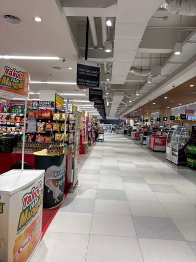 This media file captures a long, brightly lit aisle within a modern supermarket or hypermarket in Kelapa Dua, Indonesia. The view looks down the aisle towards a series of checkout counters in the distance.

The floor is tiled with alternating light grey and white squares, creating a clean, orderly appearance. On the right side of the aisle, numerous shelves are fully stocked with a variety of packaged goods, prominently snacks and food items. Large promotional displays are visible, including a cylindrical banner for "Yakult rasa Mangga" (mango-flavored Yakult) and another for "Banyalb Gaya" almond milk, both featuring "Diperkaya Vitamin D" (enriched with Vitamin D). Black hanging signs label product sections, such as "TRADITIONAL SNACK SNACK TRADISIONAL," "NUTS ANEKA KACANG" (assorted nuts), and "WAFER BISKUIT" (wafer biscuits). Other signs mention "BIRTHDAY PACKAGE" and "PROMOTION."

In the background, a series of checkout counters are numbered (e.g., 8, 9, 10). Signs like "EXPRESS COUNTER" and "SERVICE COUNTER" are visible above them. Near the checkouts, refrigerated display units hold beverages, with recognizable branding such as "Wings," "cap panda," and "Inget CINCAU." A sign pointing "TO AEON BAKER" suggests the presence of an AEON Bakery.

The ceiling features exposed white ductwork, pipes, and electrical conduits, illuminated by numerous circular and rectangular LED light fixtures. Several security cameras are mounted along the ceiling. A few blurry figures of people, likely shoppers or staff, can be seen near the checkout area, indicating typical activity in a retail environment. The use of both English and Indonesian on the signage ("OLEH OLEH INDONES," "SNACK TRADISIONAL") provides strong location cues consistent with Indonesia. The scene depicts a well-organized and functional retail space during operating hours.