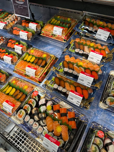 The image shows a refrigerated display case filled with various pre-packaged sushi sets. The display is organized into multiple plastic containers, each containing an assortment of sushi rolls and nigiri. Some of the sushi appears to be salmon, tuna, and other seafood. Many of the containers have red and white labels with Korean text, indicating the brand "YORIHADA". One label also says "LOTTE MART". The overall setting appears to be a grocery store or supermarket in Phu Thuy, Vietnam, given the presence of Lotte Mart. The lighting is bright, typical of a retail environment. There are no people visible in the image. The focus is entirely on the merchandise.