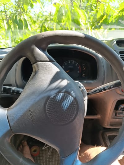 The image shows the interior of a car, focusing on the steering wheel. Through the windshield, lush green foliage is visible, suggesting an outdoor setting, possibly in Merenii Noi, Moldova. Sunlight filters through the leaves, casting dappled shadows on the steering wheel and dashboard. The steering wheel appears to be made of dark plastic and has "SRS AIRBAG" embossed on it. A wiper control stalk is visible to the right of the steering wheel. The dashboard features a speedometer and other gauges, indicating that the car is stationary. In the lower left of the frame, a bare foot and a roll of tape are visible on the floor mat. In the lower right, a person's leg and knee are visible, suggesting the driver is present. The overall scene conveys a sense of a paused moment in a vehicle parked amidst nature.