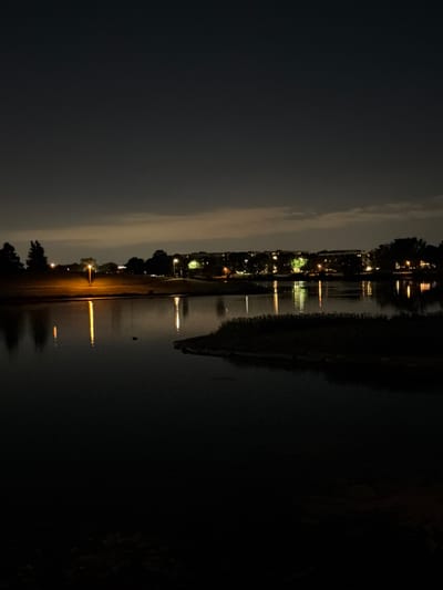 The image depicts a nighttime scene at a park in Vernon Hills, United States of America. A body of water, likely a pond or small lake, occupies the foreground and midground, reflecting the lights of the surrounding environment. In the background, a row of buildings, possibly apartment complexes or commercial establishments, is visible, illuminated by streetlights and interior lights. Trees and grassy areas are scattered along the shoreline and in the distance. The sky is dark with some cloud cover, suggesting a low-light condition typical of nighttime. There are no people visible in the image. The overall atmosphere is serene and quiet, with the lights creating an urban glow against the dark sky and water.