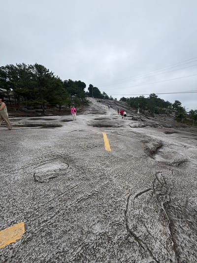 The image depicts the granite face of Stone Mountain in Stone Mountain, Georgia, United States. The foreground features the textured, gray rock surface, marked with faded yellow paint indicating a walking path. Several people are visible on the rock face at various distances; one individual is wearing a pink jacket. A line of trees grows along the upper edge of the rock. Utility wires are visible in the sky. The sky is overcast.