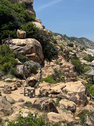 In Vinh Hai, Vietnam, a heart-shaped floral archway adorns a rocky, arid landscape. The archway, crafted from intertwined greenery and red and blue flowers, is set against a backdrop of large, weathered rocks and sparse vegetation. A small, rustic wooden table sits beneath the arch, also decorated with similar floral elements. In the distance, more rocky hills rise, dotted with patches of green foliage and several thatched-roof structures, possibly huts or shelters. The sky above is a clear, bright blue.