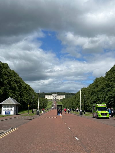 This is a photograph of the avenue leading up to Stormont Estate in Belfast City, Northern Ireland, United Kingdom. The image is taken from a low vantage point on the red-tarmac road, looking uphill towards Stormont House.

On the left side of the road, there is a small, white, hexagonal building with a pitched roof, identified by a sign as a "CHECK POINT" where "all care and caution vehicles STOP HERE." Further up the avenue on the left, a row of tall, mature trees lines the grassy verge.

The road itself is wide and lined with yellow and white markings. Several people are walking along the avenue in both directions. There are also some orange traffic cones visible in the middle of the road.

On the right side of the road, a bright green ice cream van with "SOFT ICE CREAM" written on it is parked, with several people standing near it. More trees line the right side of the avenue, with some people walking in the background.

In the distance, Stormont House is prominently visible at the top of a wide, manicured lawn. The building is large and imposing, with a symmetrical facade and a flagpole on its roof. The sky above is a mix of dramatic, dark grey clouds and patches of blue sky. The overall scene suggests a public space with visitors enjoying the grounds.