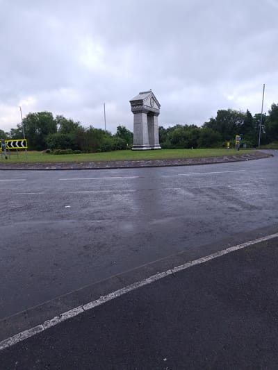 The image depicts a view of a roundabout with a prominent monument in the center. The monument is a tall, grey, classical-style structure with a clock face at the top. The roundabout is surrounded by green grass and trees, suggesting a suburban or parkland setting. The road surface is wet, indicating recent rainfall, and there are lane markings visible on the asphalt. Traffic signs are present on the left side of the roundabout, including a yellow directional sign with black chevron markings. The sky is overcast with grey clouds. The scene appears to be an outdoor public space, likely a junction within the city of Newport, United Kingdom.