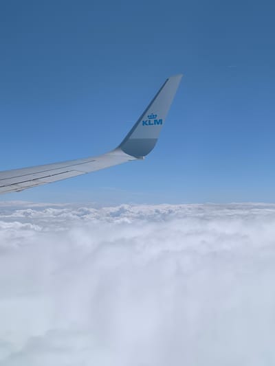 The image shows a view from an airplane window. A KLM airplane wing is visible in the upper left of the frame. Below the wing, a dense layer of clouds stretches across the middle and bottom portion of the picture. Above the wing, a clear blue sky is present. High in the sky, a contrail from another airplane can be seen. There is no indication of the city of Fischamend, Austria.