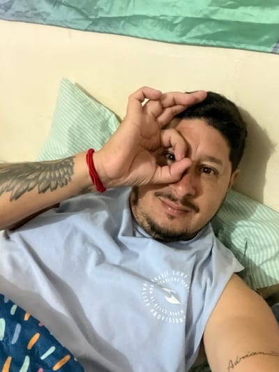 A male subject, estimated to be in his 30s-40s, is depicted indoors, likely in a bedroom in Bernal Este, Argentina. He is shown from the chest up, lying on his back, directly facing the viewer. His right hand is raised, with the thumb and forefinger forming an "OK" gesture around his right eye. He has dark hair, a trimmed beard and mustache, and brown eyes. The subject is wearing a light blue sleeveless t-shirt with a white circular graphic logo reading "BREEZE SURF ESTABLISHED SIXTY NINE QUALITY AT BELLS BEACH PROVISION." A red string bracelet is visible on his left wrist. A grey-toned wing tattoo is present on his upper left arm, and a tattoo in script, possibly "Adriana," is visible on his right bicep area. The background features a cream-colored surface, a light green striped pillow, and a draped green fabric. A section of a dark blue blanket with abstract light blue, orange, and white shapes is visible in the lower left corner.