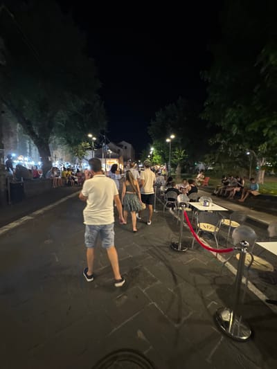 The image depicts a nighttime scene on a cobblestone street in Orosei, Italy. The street is lined with trees and buildings, with an outdoor seating area for a restaurant or cafe on the right side. Several people are visible, some walking and others seated at tables. The foreground shows the back of a man wearing a white polo shirt and blue shorts, walking away from the camera. To his right, a woman in a patterned skirt and top walks in the same direction. Further down the street, more people are gathered, some sitting at tables in the restaurant's outdoor seating area, and others standing or walking along the sidewalk. The lighting suggests it is evening, with streetlights and lights from buildings illuminating the scene. The overall atmosphere appears to be one of a pleasant summer evening, with people enjoying the outdoors.