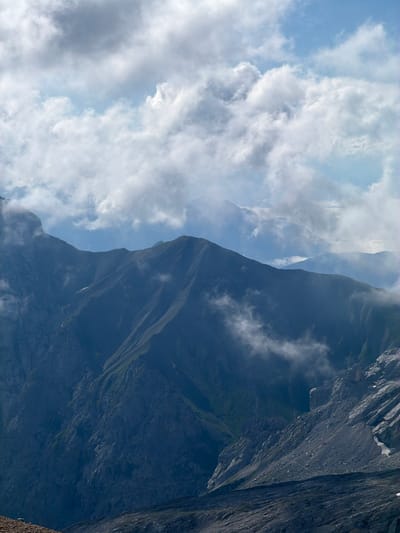 This is a vertical, full shot of a mountain landscape under a partly cloudy sky. The foreground is dominated by the dark, rocky slopes of a mountain range, with some patches of green vegetation visible in the crevices. Wisps of cloud drift around the peaks, partially obscuring their rugged features. The middle ground features more jagged mountain ridges, also shrouded in mist and clouds. In the distance, further mountain ranges fade into a hazy blue. The sky above is a mix of bright blue patches and soft, white clouds, some of which appear to be illuminated by sunlight. The overall impression is one of a vast, wild, and somewhat dramatic mountain environment, likely captured in Austria, possibly near Wildermieming, known for its alpine scenery. There are no people or specific man-made objects visible, emphasizing the natural grandeur of the scene.