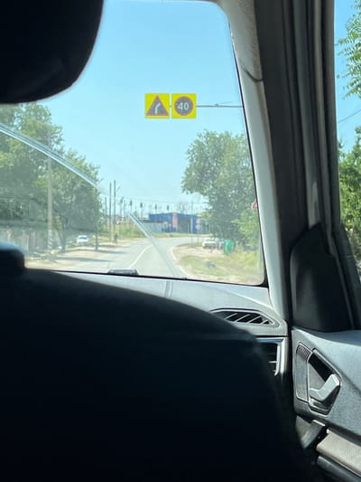 The image captures a view through a car windshield, likely taken from the front passenger seat, looking out at a sunny day on a road in the village of Ilyinka, Russia. Two traffic signs are visible on the windshield. The sign on the left is a yellow triangle with a black curved arrow pointing right, indicating a turn ahead. The sign on the right is a yellow circle with a red border and the number "40" in black, signifying a speed limit of 40 kilometers per hour.
The road ahead curves to the right, with trees lining both sides. In the middle distance, several utility poles are visible, and a few buildings, including one with blue siding, are present. A white car is parked on the left side of the road, and a person on a bicycle is visible further down the road on the right. The foreground of the image is dominated by the dark interior of the car, with a black headrest or seat visible and part of the car's dashboard. The overall impression is of a clear, warm day, with normal daily activity on a road in a rural or semi-rural area.