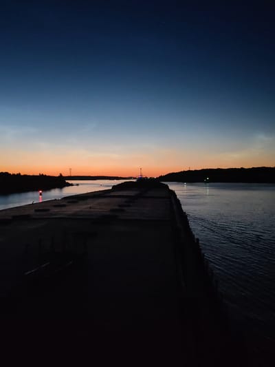 The image captures a twilight scene over a body of water, possibly a canal or river, with a concrete embankment in the foreground. The sky transitions from a deep blue at the top to a vibrant orange and yellow near the horizon, indicating the sun has recently set. Silhouetted trees line the distant shores. A red navigation light is visible on the left side of the water, and a few other lights, some appearing green, are scattered further down the waterway. The concrete embankment is dark and textured, with some debris or structures faintly visible on its surface. The water itself shows gentle ripples and reflects the fading light. The location is identified as Sud'bitsy, Russia.
