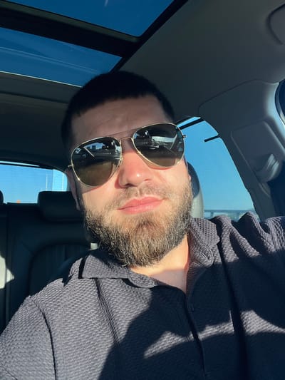 A close-up, selfie-style image shows a man with a beard and sunglasses inside a car. He is wearing a dark, textured polo shirt. The car's sunroof is open, revealing a bright blue sky with some wispy clouds. The interior of the car is visible, including the black leather seats and the dashboard. The lighting suggests it is daytime, with sunlight casting shadows. The reflection in his sunglasses shows buildings and a road, indicating an urban or suburban environment. There is no visual information in the image that allows for the specific location of Florești, Romania to be identified.