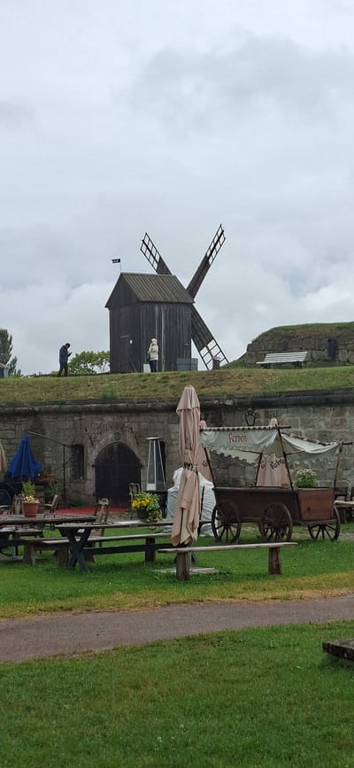 The image depicts an outdoor scene with a prominent windmill in the background, suggesting a historical or rural setting. In the foreground, there are several wooden tables and chairs, with some umbrellas set up, indicating an outdoor seating area, possibly for a cafe or restaurant. A cart with wheels and a canopy is also visible, which could be a food stall or part of the establishment's decor. The ground is covered with green grass, and a pathway runs alongside it. The sky is overcast, hinting at a cloudy or possibly rainy day.
There are two people visible in the image. One person, wearing a light-colored jacket and pants, stands near the base of the windmill. Another person, dressed in a blue denim jacket, is standing further away, looking down at what appears to be a phone. The overall atmosphere seems calm and perhaps a bit subdued due to the weather.
The windmill itself is made of dark wood and has large sails, two of which are partially visible. A small black flag with white text is attached to the windmill. The stone wall of a building is visible behind the tables and chairs, with a dark arched doorway. There is some greenery, including potted plants with yellow flowers, arranged around the seating area.
Although the image is rotated, the context provided by the location cue (Kuressaare, Estonia) and the visual elements like the windmill and stone architecture suggest a historical site or a tourist attraction in that area. The text on the flag on the windmill is partially visible but unreadable. The text on the canopy of the cart appears to read "fenn" which could be part of a brand name or advertisement.