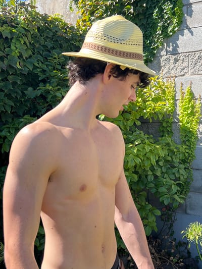 The image, taken in Quilmes Oeste, Argentina, captures a male subject from the chest up, positioned outdoors against a backdrop of green foliage. The individual, appearing to be a young adult with fair skin and dark, curly hair, is shirtless and wears a light-colored, possibly straw, fedora-style hat with a brown patterned band. He is viewed in profile, facing right, with his head slightly tilted down, suggesting a downward gaze. His left shoulder and arm are visible, along with his chest and abdomen. The background features a light grey concrete block wall mostly covered by dense, vibrant green climbing plants, likely ivy. Bright sunlight illuminates the scene, creating distinct shadows on the wall and highlighting the subject's skin. A partial view of a dark garment, possibly swimwear, is visible at the bottom edge of the frame. In the lower right background, an out-of-focus spherical green plant cluster with thin stems is present.