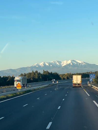 This image depicts a multi-lane highway scene under a clear blue sky, with a range of snow-capped mountains visible in the distant background. The vantage point is from inside a vehicle traveling forward. The location is identified as the vicinity of Salses-le-Château, France, by a blue highway sign on the right shoulder which reads "Aire de Salses-le-Château 1000m" and includes a parking symbol.
The highway consists of at least two lanes of travel in the foreground, marked by white dashed lines, and is separated by a median strip containing a metal barrier. In the left lane, ahead of the camera's position, a white semi-truck with a silver trailer is observed, featuring an orange triangular warning sign on its front bumper. Further ahead in the right lane, a white box truck is visible from its rear. A smaller white vehicle, possibly a car or van, is present in the distant left lane. A small yellow triangular sign on a metal pole is positioned within the median strip. Beyond the median, a dense line of green trees borders the highway, separating it from the mountainous horizon. A reddish-brown building or structure is faintly discernible above the treeline on the right side. The scene is brightly and evenly illuminated by natural daylight. No individuals are explicitly visible.