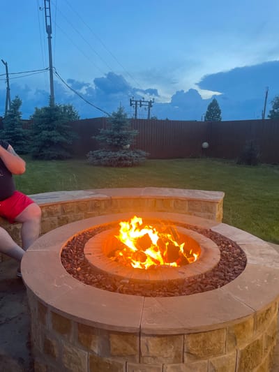 A backyard scene in Chulym, Russia, features a stone fire pit with a roaring fire, surrounded by small, dark rocks. A person wearing red shorts and dark clothing sits on a curved stone bench next to the fire pit. Beyond the fire pit, a lush green lawn is visible, bordered by a dark wooden fence. Several evergreen trees are planted in the yard, and in the background, power lines and utility poles stretch across a cloudy, blue twilight sky. A pale spherical object hangs on the fence in the middle distance.