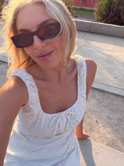 The media file is a selfie of a young woman with blonde hair and sunglasses. She is wearing a white, eyelet fabric dress with a ruffled neckline. The woman is outdoors, seated on concrete steps or a bench. The lighting suggests it is late afternoon or early evening, with warm sunlight casting shadows. In the background, there are hints of greenery, possibly palm trees, and a red brick wall with graffiti. The overall scene appears to be a casual, sunny day in an urban or park-like setting. The woman's expression is relaxed and she is looking towards the camera. There is visible text on the side of her sunglasses, which reads "VOGUE". The location context is identified as Benimàmet-beniferri, Spain.