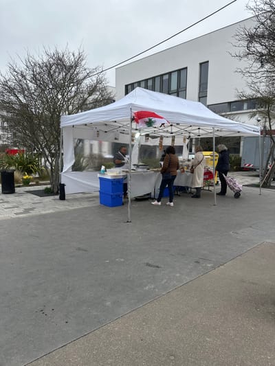 The image displays an outdoor market scene located in Serris, France, under an overcast sky. A white pop-up canopy tent, functioning as a market stall, is centrally positioned on a grey paved surface. A Lebanese flag, characterized by red stripes and a green cedar tree on a white field, is affixed to the front-right support of the canopy.

Within the stall, a male vendor with dark hair and a beard, wearing a grey short-sleeved shirt, stands behind a light-colored table. Various food items, including what appear to be pastries or savory dishes, are displayed on the table. Two stacked blue plastic crates are situated at the front-left base of the stall.

Three female customers are present at the stall, facing the vendor and the displayed items. From left to right, the first woman wears a brown quilted jacket and blue jeans. The second woman wears a long, light-colored (cream) coat. The third woman, identifiable as older with grey hair, wears a dark jacket and dark trousers, and holds a purple floral-patterned shopping trolley.

The background on the left features a section of landscaping with bare deciduous trees, green shrubbery, and patches of yellow flowers (likely daffodils). A black cylindrical trash receptacle stands on the far left. Behind the stall, a modern white building with dark rectangular windows is visible. To the far right, a portion of a white utility vehicle and a yellow bin are partially obscured. Overhead utility lines are faintly visible against the sky. The ground is a uniform grey pavement.