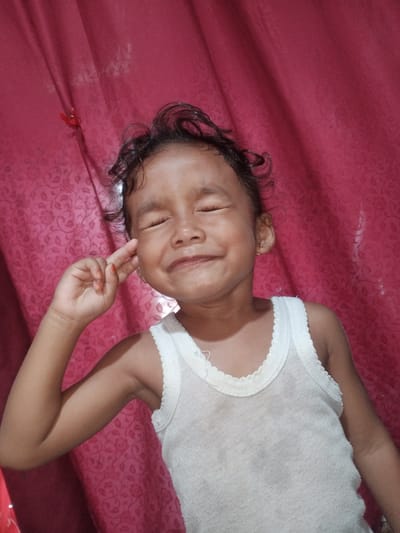 A young child with dark curly hair is shown from the chest up, posing with their eyes closed and a finger held to their temple, against a red curtain background with a floral pattern. The child is wearing a white tank top. The image appears to be a casual photograph taken indoors. There is no discernible information that places the image in Cotseutui, Indonesia.