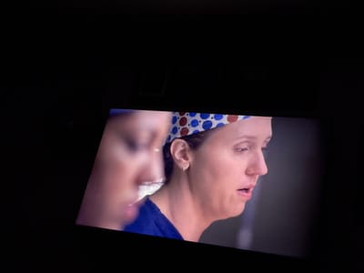 The image displays a screen showing two women in a dark environment. The primary subject, a woman in the foreground on the right, is shown in profile facing left. She has fair skin, light brown hair, and wears a blue scrub top and a white scrub cap patterned with blue and reddish-brown polka dots. Her mouth is slightly open, and her eyes appear to be cast downwards, conveying an expression of distress or deep concentration. A small silver hoop earring is visible in her left ear. A second woman, heavily blurred and out of focus, is partially visible on the left side of the screen, also in profile. The background within the screen's display is a dark, nondescript gray-blue. The overall scene suggests a moment from a medical drama or similar program being viewed in a dimly lit room. The specific location in Casa Blanca, Mexico, is not discernible from the visual content of the image.