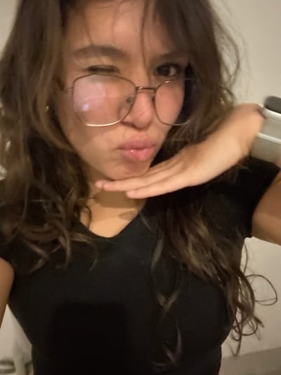 A close-up, chest-up image depicts a female subject with long, wavy brown hair. She is wearing large, gold-framed eyeglasses. Her left eye is closed in a wink, and her lips are pursed. Her right hand is positioned beneath her chin, with the palm facing upwards. She is dressed in a black V-neck top. A smartwatch with a light-colored band is visible on her left wrist. The background is an unfocused, light-colored surface. The image contains no visual information to confirm the stated location of Casa Blanca, Mexico.