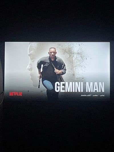 This image depicts a photograph of an illuminated screen displaying the movie poster for "Gemini Man," captured in a dark room within the city of Ksar-el-Kebir, Morocco. The poster features a prominent depiction of Will Smith in the foreground, middle-aged, running towards the right. He is dressed in a dark collared shirt and blue jeans, holding a black pistol in his left hand, and has a dark strap or bag slung over his right shoulder. Behind him, a translucent, younger profile of Will Smith faces left. The background of the poster transitions from a light gray on the left to white on the right, with the right side showing a fragmented or dissolving texture. Overlaid text includes "GEMINI MAN" in large white capital letters, "NETFLIX" in red in the bottom left corner, and Arabic text (أعداء. أكشن, خشونة) directly beneath "GEMINI MAN." The screen is centrally framed by the dark, unidentifiable surroundings of the room where the photograph was taken.