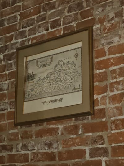 A framed map of Kentucky hangs on a brick wall. The map depicts various towns, cities, and geographical features of the state, with labels for neighboring states like Indiana, Illinois, and Tennessee. There are small illustrations of people and horses on the map. The map is housed in a golden frame with a beige matting. The brick wall has a rustic texture with varying shades of red and brown bricks and off-white mortar. The lighting suggests an indoor setting. The map appears to be a historical or decorative piece. The image does not provide information about a specific location in Shelbyville, United States of America.