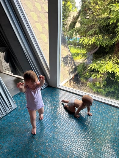 Two infants are on a tiled floor indoors near a window. One infant wears a light purple shirt and is lying on their stomach. The other infant wears a diaper and is on their hands and knees. Outside the window, there are trees and a building with a tan roof. The specific location in the city of Чёрное, Russia is impossible to ascertain from the visual information.
