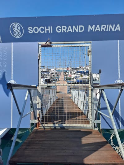 This image shows a bright, sunny day at a marina, likely in Chvizhepse, Russia, as indicated by the text on the sign. The main subjects are the yachts moored in the marina, visible through a metal gate. The gate itself is a prominent object, made of wire mesh and metal framing, and appears to be a part of a walkway or pier.

To the left of the gate, a large blue sign displays white text vertically. The text reads "SOCHI GRAND MARINA" along with a circular logo featuring an intricate knot design. Further down the sign, though partially obscured and rotated, are coordinates that appear to be "43° 34' 30.9" N" and "39° 43' 07.0" E".

The scene also includes a wooden pier or walkway on the right side of the image, casting a strong shadow. Metal railings and structures are present, suggesting a well-maintained facility. The water in the marina is a dark green, reflecting the sky and the surrounding structures. The sky itself is a clear, bright blue, indicating good weather. There are no people clearly visible in the frame, and the focus is on the marina's infrastructure and the boats. The overall impression is one of a peaceful and functional marine environment.