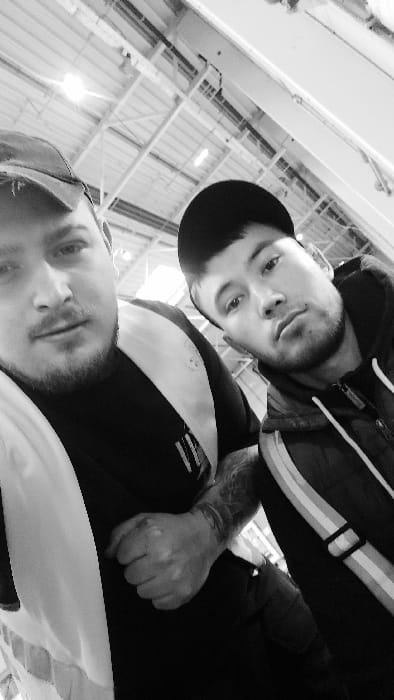 A black and white selfie image depicts two adult males within an indoor industrial or warehouse environment, located in the city of Senec, Slovakia.
The man positioned on the left side of the frame, closer to the camera, wears a dark baseball cap, a black t-shirt, and a partially visible light-colored reflective safety vest over the t-shirt. He has a full beard and mustache. His right fist is clenched, revealing tattoos on the back of his hand and forearm.
The man on the right wears a dark baseball cap and a dark hooded jacket with light-colored vertical stripes on the upper arm. He has short facial hair.
Both individuals are looking directly forward, toward the camera.
The background features a high industrial ceiling structure, composed of white metal beams and panels. Multiple overhead lighting fixtures are visible, one of which emits a distinct glare. Dark linear elements, possibly indicative of overhead tracks or ventilation systems, are also present. The environment appears brightly lit.