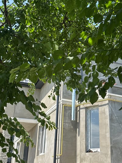 The media file captures a view looking upwards towards a building facade, partially obscured by the lush green foliage of a tree. The scene is set outdoors, likely in a residential or urban area given the building's appearance.

The building features a concrete wall with a long, narrow window on the upper level. The window frame is white and has a sticker with text on it. Below this window, a silver cylindrical pipe runs horizontally, possibly part of a ventilation system. Further down the wall, a bright yellow pipe is visible, also running horizontally. The building's architecture suggests it might be an apartment building or a similar structure.

The tree's branches and numerous green leaves dominate the foreground and midground of the image, creating a natural contrast with the man-made structure. Sunlight filters through the leaves, casting dappled shadows and highlighting the vibrant green of the foliage. The sky is visible in patches through the leaves, suggesting it is a clear day.

The time of day appears to be daytime, likely morning or afternoon, given the strong sunlight and the shadows cast by the building. The weather seems fair and sunny. There are no specific events or activities depicted, rather it's a static observation of the environment. The overall mood is peaceful and natural, with the greenery softening the harshness of the concrete building. The sticker on the window appears to have repeated text, possibly brand names or installation instructions, but it's difficult to read clearly. The location context of Yablunivka, Ukraine, places this scene within a specific geographical area.