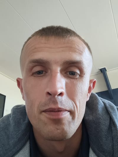 The image is a close-up selfie of a white male with a buzz cut, taken indoors. He is wearing a gray hooded jacket. His face is the primary subject, with his eyes open and looking towards the camera. The background appears to be a ceiling with light fixtures and a wall with some dark blue elements. There is no visible indication of the city of Tinte, Netherlands, in the image.